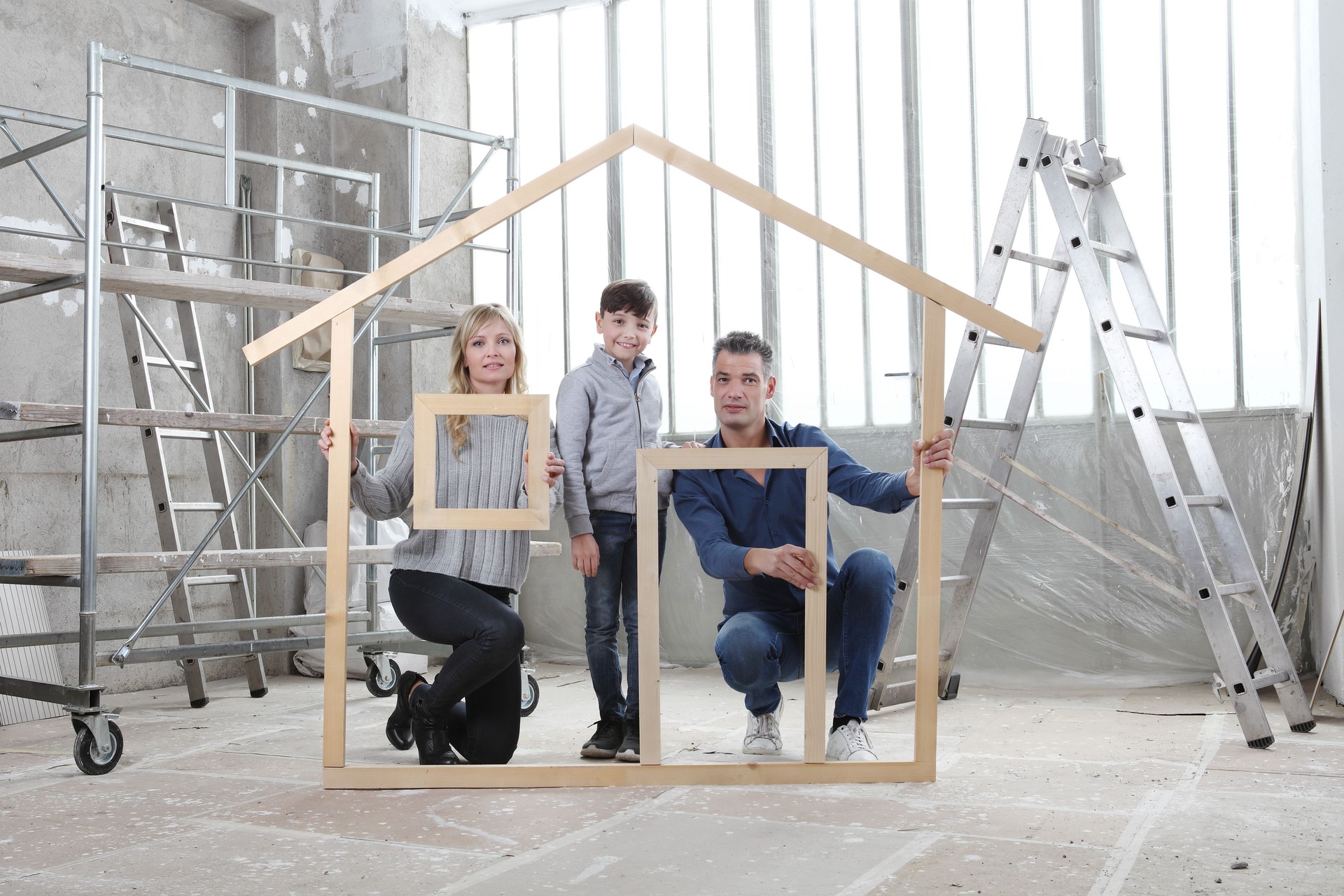 New building residential house purchase apartment concept. Happy family with child hold with their hands the wooden house figure frame with door, window, wall and roof. Inside of interior construction site